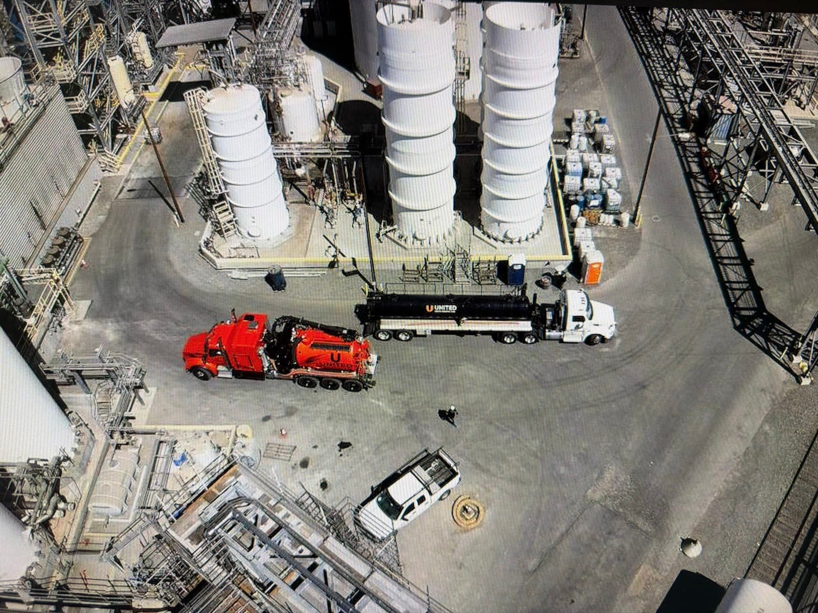 Aerial view of a vacuum truck and a flatbed truck working inside an industrial plant, between storage silos and pipe racks