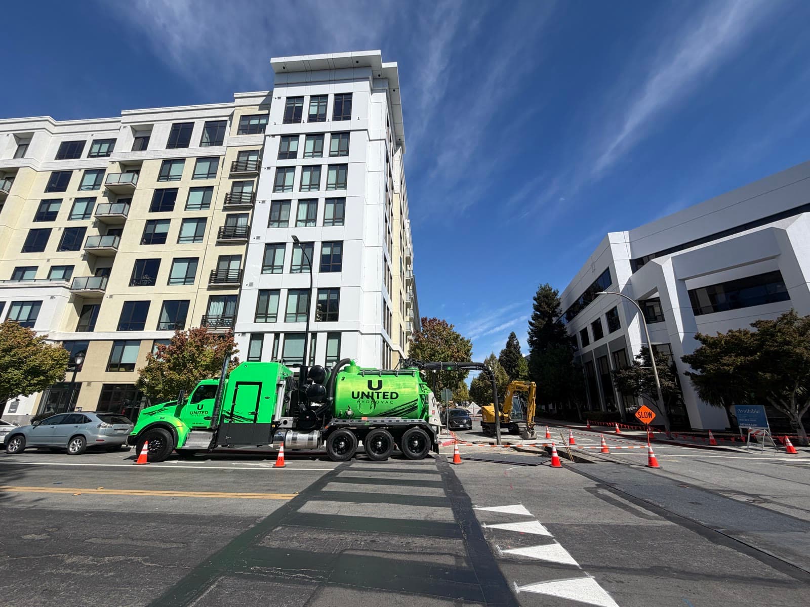 Wide-angle view of a green United Hydrovac truck and a mini excavator at a coned-off street job between an apartment building and an office building